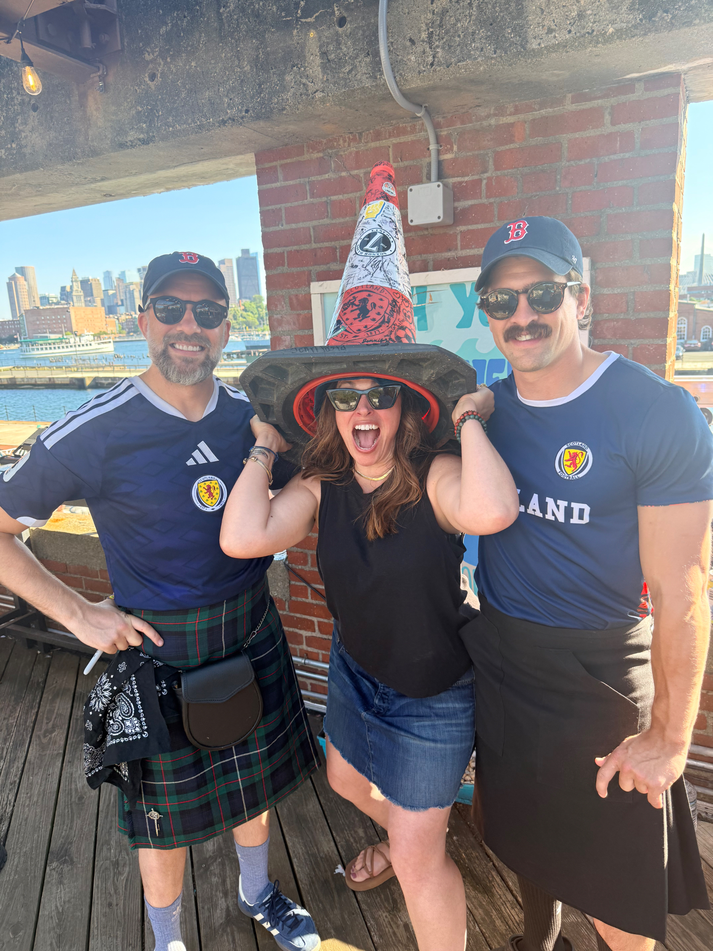 Reed in a traffic cone hat between two friends in Scotland jerseys and a kilt, Boston waterfront